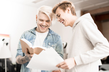 Obraz premium Caucasian hipster-like tattooed male student and his friend looking at pictures in textbook and smiling joyfully