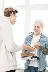 Obraz premium Tattooed adult student with dyed blond hair exchanging books with his young college mate and smiling happily, low angle view