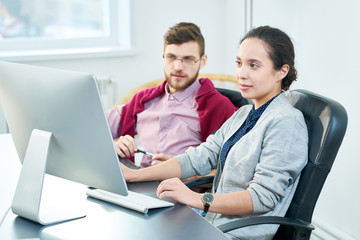Young female office worker looking at computer screen attentively while sitting at office table with bearded male colleague