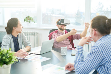 Young Caucasian office workers smiling cheerfully while looking at their colleague having fun in virtual reality headset