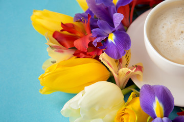 a plan of a white forfor-cup with a saucer and coffee around which lie tulips, narcissus, and orchids that lie on a blue background