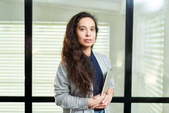 Waist-up portrait of young Caucasian woman with long dark hair standing in office with digital tablet and looking at camera