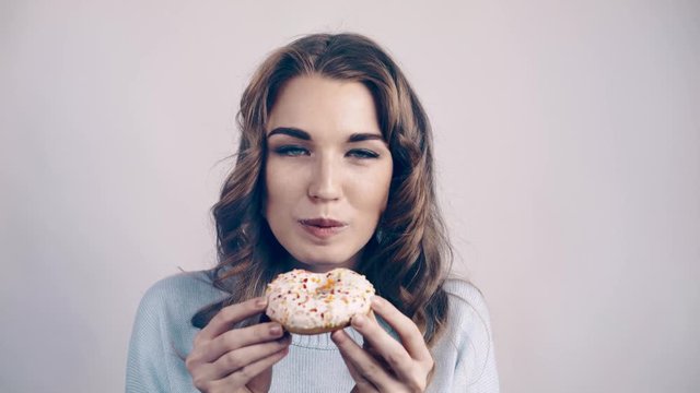 Pretty young woman with fair hair wearing a sweater is eating a tasty doughnut. Locked down real time close up shot