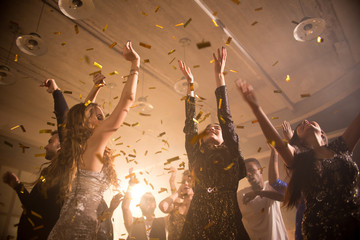 Cheerful group of young men and women dancing with raised hands while having fun at trendy night club, golden confetti flying in air