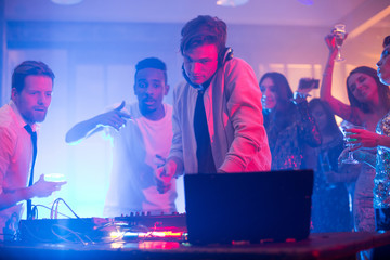 Talented young DJ mixing music with professional equipment while group of cheerful friends dancing...