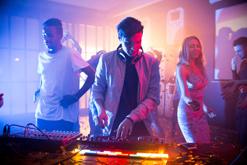 Confident young DJ playing trendy music with help of mixer while cheerful friends dancing behind...