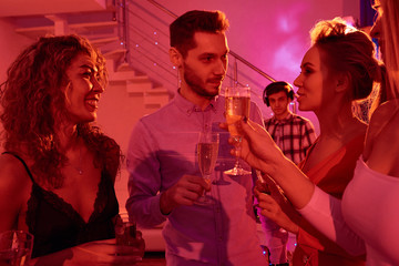 Waist up portrait of handsome man surrounded by beautiful young women and chatting happily while drinking champagne during house party in red light