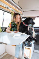 happy young woman reading a book and playing with her dog in a camper van