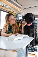 happy young woman reading a book and playing with her dog in a camper van