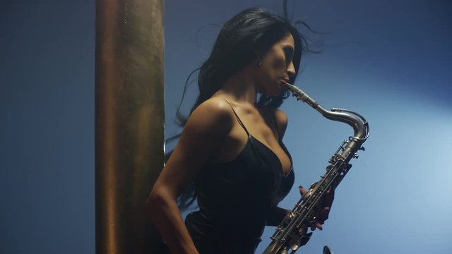 Young, beautiful, sexy, brunette woman dancing and playing golden saxophone on dark background . Shot on RED HELIUM Cinema Camera in slow motion. 