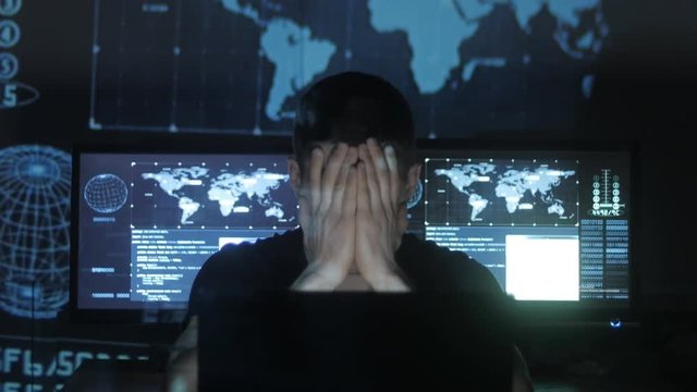 Young Man geek hacker overworking at computer and suffers from a headache in cyber security center filled with display screens.