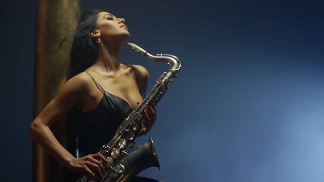Young, beautiful, sexy, brunette woman dancing and playing golden saxophone on dark background . Shot on RED HELIUM Cinema Camera in slow motion.