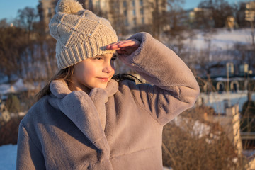 beautiful girl stands in a gray fur coat and a knitted beige hat on the background of houses