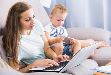 Satisfied woman is productively working behind laptop while child playing on tablet