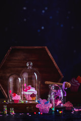 Colorful cupcakes papercraft under a glass dome. Ideal confectionery concept with copy space. Dark food photography with confetti and sprinkles.