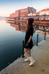 Young woman in hat and cute summer dress standing on the pier with peaceful town scenery, looking at sunset