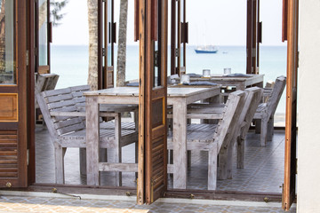 Table and wooden chairs in empty cafe next to the sea water on the beach, Thailand