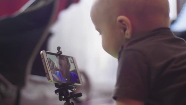 Small baby sitting on the floor communicates on video call in a smartphone with his caucasian father and touches the screen. Indoor 4K left side view closeup footage.