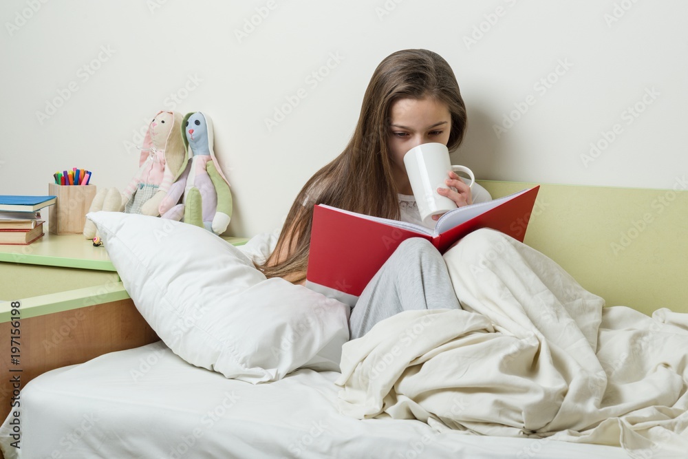 Wall mural Teen girl 10 years old in pajamas reads book and holds cup of tea.