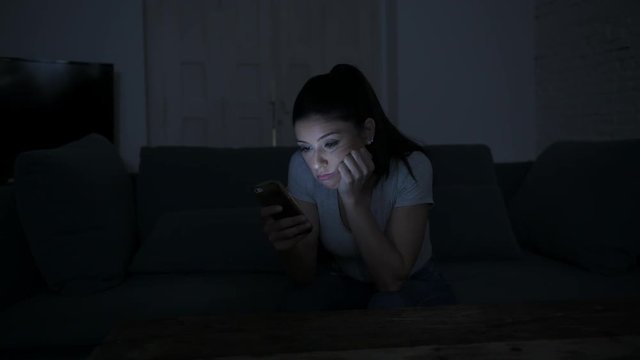 young attractive and tired hispanic 30s woman with mobile phone looking serious and bored using dating app sitting at apartment living room couch late night