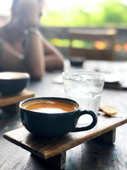 A cup of coffee with a glass of cold water, stands on a wooden stand and a wooden table in a cafe. In the background sat a man.