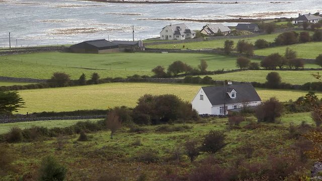 Still shot of green Irish hills and houses. Hedgerows and stone walls divide the properties with a body of water in the background.