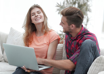 young couple is using a laptop and smiling while sitting on sofa at home