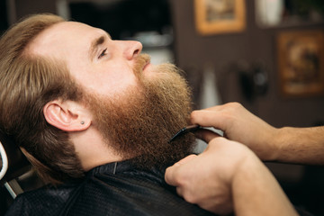 Obraz premium Bearded man getting a haircut by a professional hairdresser using comb and grooming scissors. Closeup view with shallow depth of field