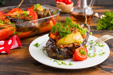 Eggplants stuffed with minced meat and cooked with fresh tomatoes and bell peppers on white plate and in baking dish on wooden rustic table.