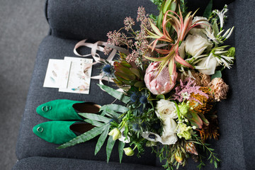 Rich green wedding bouquet with pink ribbons on the grey armchair. Green bridal shoes, and a wedding complimentary lying near the bridal lush bouquet. Details