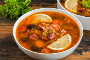 Thick and hearty Russian tomato soup Solyanka with turkey, sausages, pickled cucumbers, black olives and fresh lemon in white casserole and bowl on the wooden rustic table.