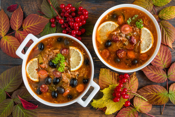 Thick and hearty Russian tomato soup Solyanka with turkey, sausages, pickled cucumbers, black olives and fresh lemon in white casserole and bowl on the wooden rustic table, top view.