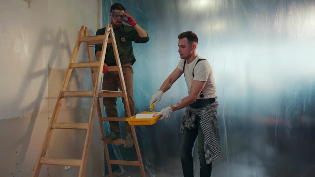 Young man in a dirty uniform helps his co-worker on a ladder, gives him the paint roller, then goes to freshly painted wall to check the result. Working hard, partnership. Slow motion, camera