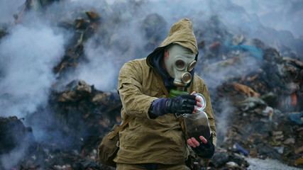 a man in a gas mask holds a sprout in the flask, against the background of fuming debris