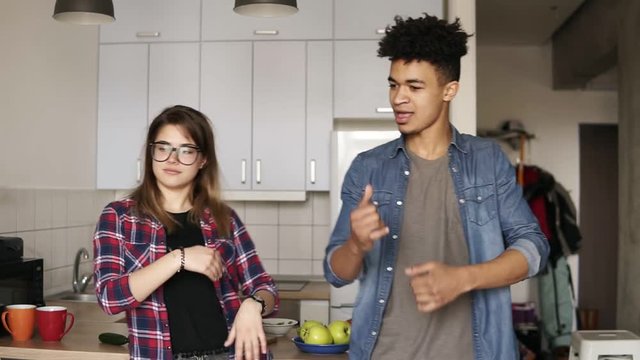 Attractive caucasian girl with stylish hipster glasses and playful guy, both in ther 20's, dancing in the kitchen, enjoying youth, having fut time together.