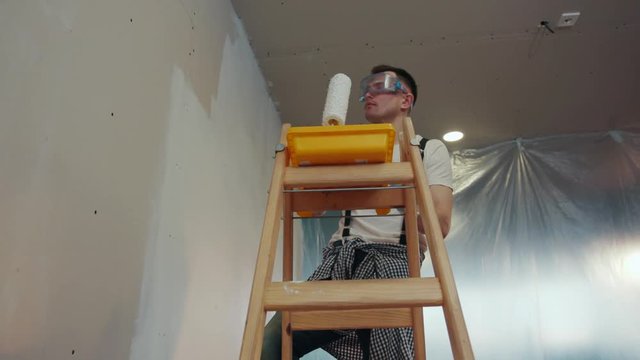A handsome, skilled house painter in protecting gloves and goggles stands on the ladder, uses the painting trail and roller to paint the wall in white color. Western style renovation. Slow motion