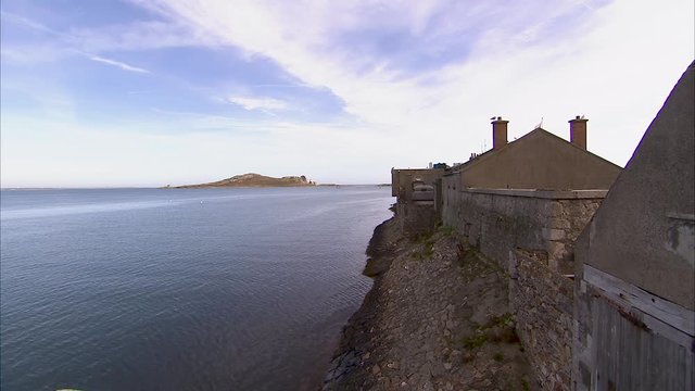 Still shot of a rocky shoreline of an old Irish town made of stone and wood. An island can be seen in the distance, under the blue cloudy sky.