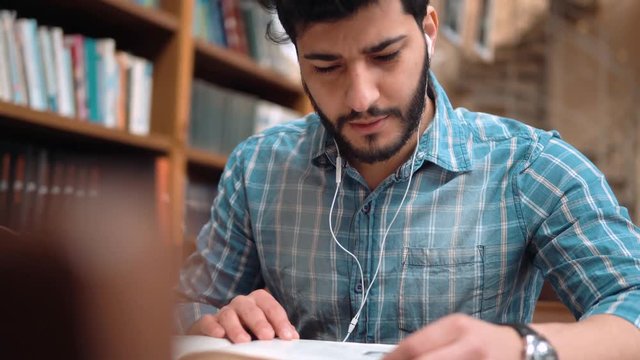 Focused student mumbling text at brown desk in the library, handsome black-haired man turning book page as listening to music