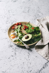 Vegetarian sandwiches with avocado, poached egg, spinach, cherry tomatoes on whole grain toast bread on ceramic plate with textile napkin over white marble background. Copy space. Toned image