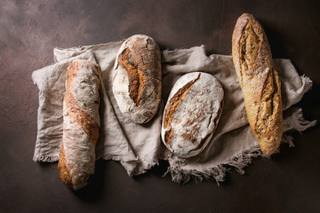 Variety of loafs fresh baked artisan rye and whole grain bread on linen cloth over dark brown...