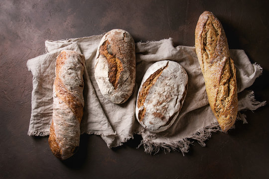 Variety of loafs fresh baked artisan rye and whole grain bread on linen cloth over dark brown texture background. Top view, copy space.