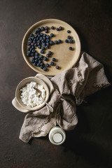 Ceramic bowl of homemade cottage cheese served with blueberries and bottle of milk on linen cloth over dark brown texture background. Top view, space