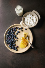 Ceramic bowl of homemade cottage cheese served with blueberries, bottle of milk and honeycombs over dark brown texture background. Top view, space
