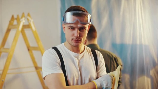 Sexy young house painter with a stain of white paint on his forehead holds the rendering trowel, crosses his arms on his chest and seductively looks straight to camera. Passion, romantic atmosphere