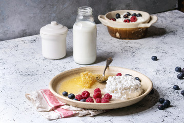 Ceramic plate of homemade cottage cheese served with blueberries, raspberries, bottle of milk and honeycombs over white marble texture table as background.