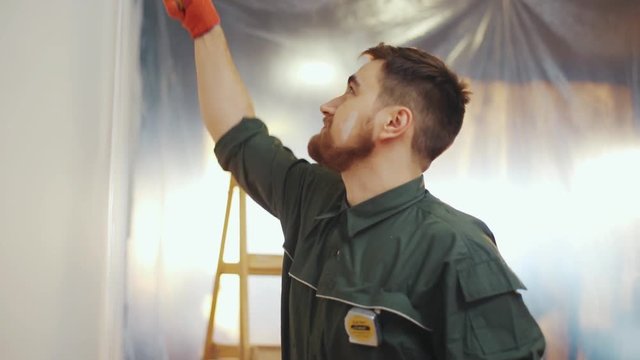 Young bearded man paints the wall, then turns to his friend, tells him a joke, both are happily laughing. Positive emotions, good mood, having fun. Slow motion, camera stabilizer shot