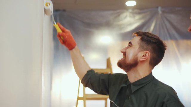 Cheerful bearded house painter paints the wall with the paint roller, then turns to his friend, tells him a joke, gestures actively, dances, both laugh happily. True friends, male friendship, happy