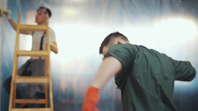 Handsome young man decides to have a break from the refreshment process, hilariously moves with the paint roller, his co-worker joins him. Party hard, positive mood, having fun. Slow motion, camera