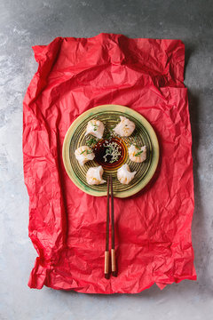 Asian steam potstickers dumplings stuffed by shrimps, served on ceramic plate with soy sesame sauce and chopsticks on crumpled red paper over grey texture background. Top view, space.
