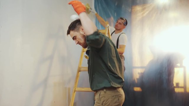 Handsome young builder decides to have a break from the refreshment process, hilariously dances, juggles the paint roller, his co-worker applauses him. Party hard, positive mood, having fun. Slow